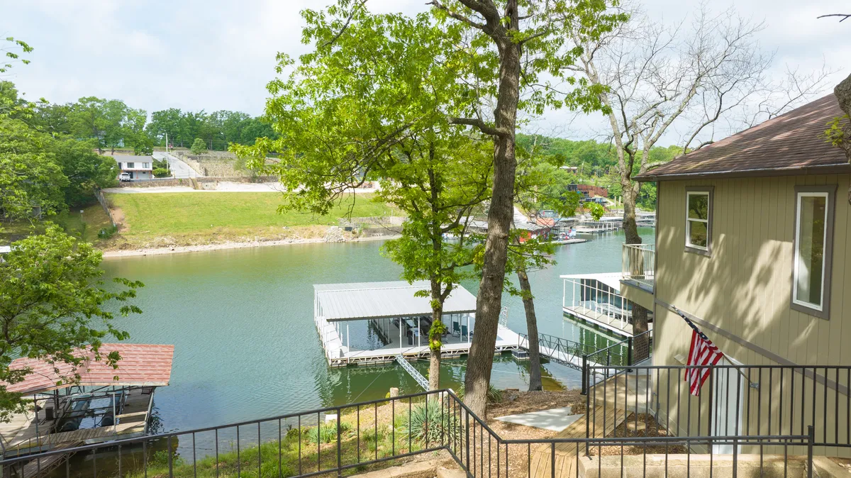 A serene lakeside view featuring calm water, private docks, lush green trees, and a glimpse of a house nestled along the shoreline. The scene evokes a sense of tranquility and easy access to water activities, typical of Osage Beach's recreational appeal.