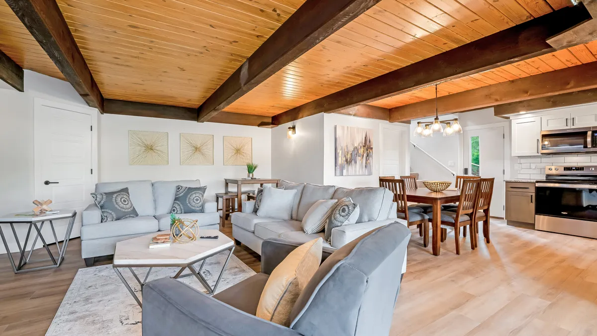 A spacious, cozy living and dining area featuring plush sofas, a beautifully set wooden dining table, stunning wall artwork, and exposed wood beams. The space is arranged for social gatherings, family meals, and relaxed lounging, radiating comfort and togetherness.