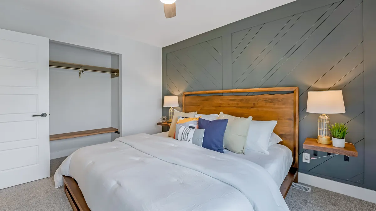 A cozy bedroom with a wooden headboard, modern bedside lamps, decorative pillows, and a striking geometric accent wall. The setting feels tranquil and restful, ideal for unwinding or curling up with a book.