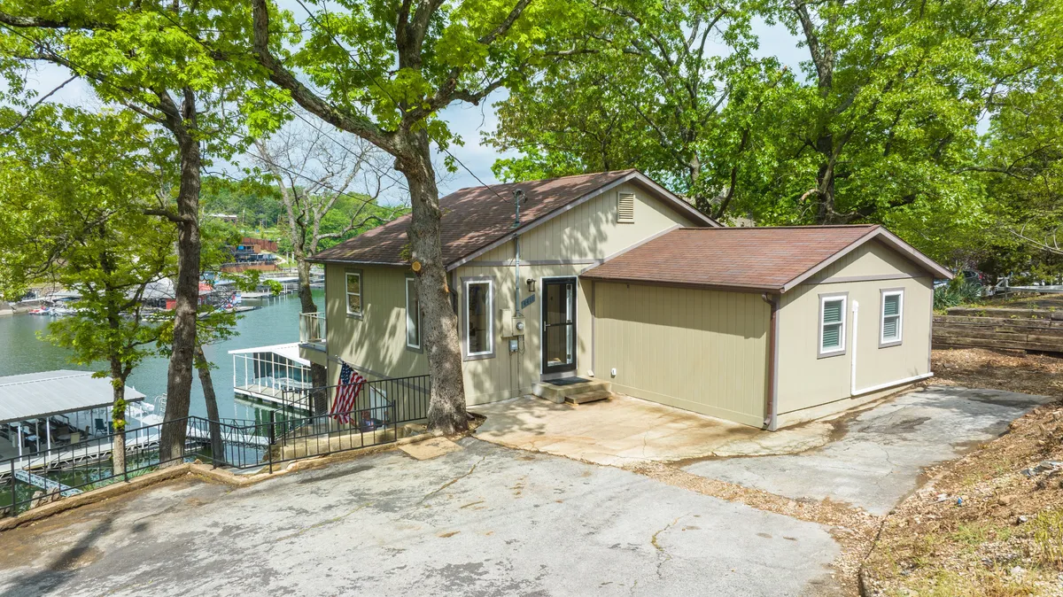 The image shows the exterior of the house nestled among mature trees, with a view of the tranquil lake just beyond. The scene exudes peace and offers a sense of spaciousness, ideal for outdoor walks and activities with pets. The lush green surroundings and water view emphasize the property's connection to nature.