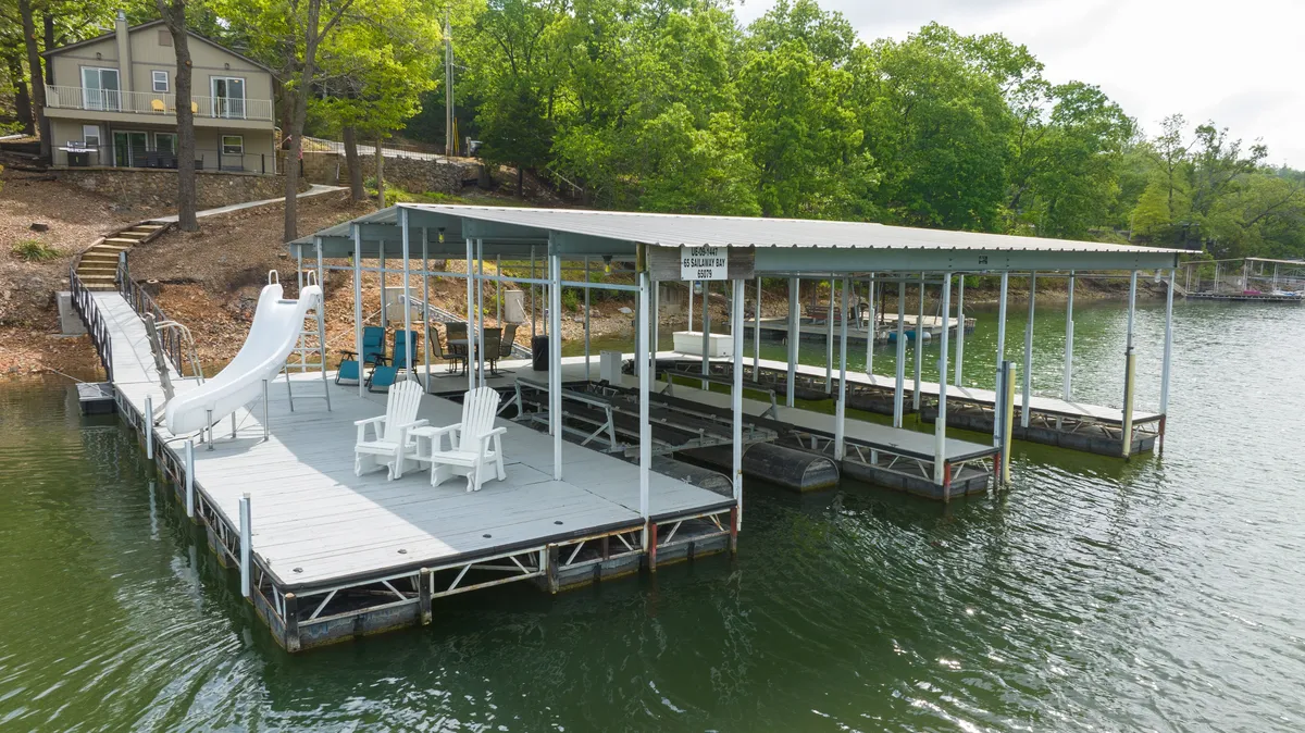 The image showcases a serene lakefront setting with a spacious dock, colorful Adirondack chairs arranged for relaxation, a fun water slide leading into the lake, and lush trees bordering the shoreline. The golden hues of sunset reflect off the water, inviting guests to enjoy swimming, boating, and making memories outdoors.