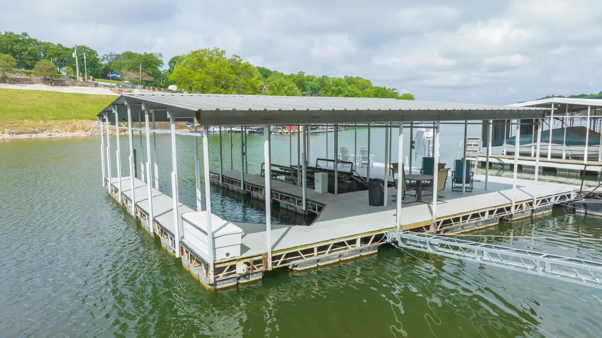 A serene dock stretches out over calm waters, surrounded by lush greenery. The open space on the dock is perfect for fishing, launching watercraft, relaxing, or gathering with friends and family. The setting sun adds a warm, golden glow to the scene, enhancing its tranquil appeal.