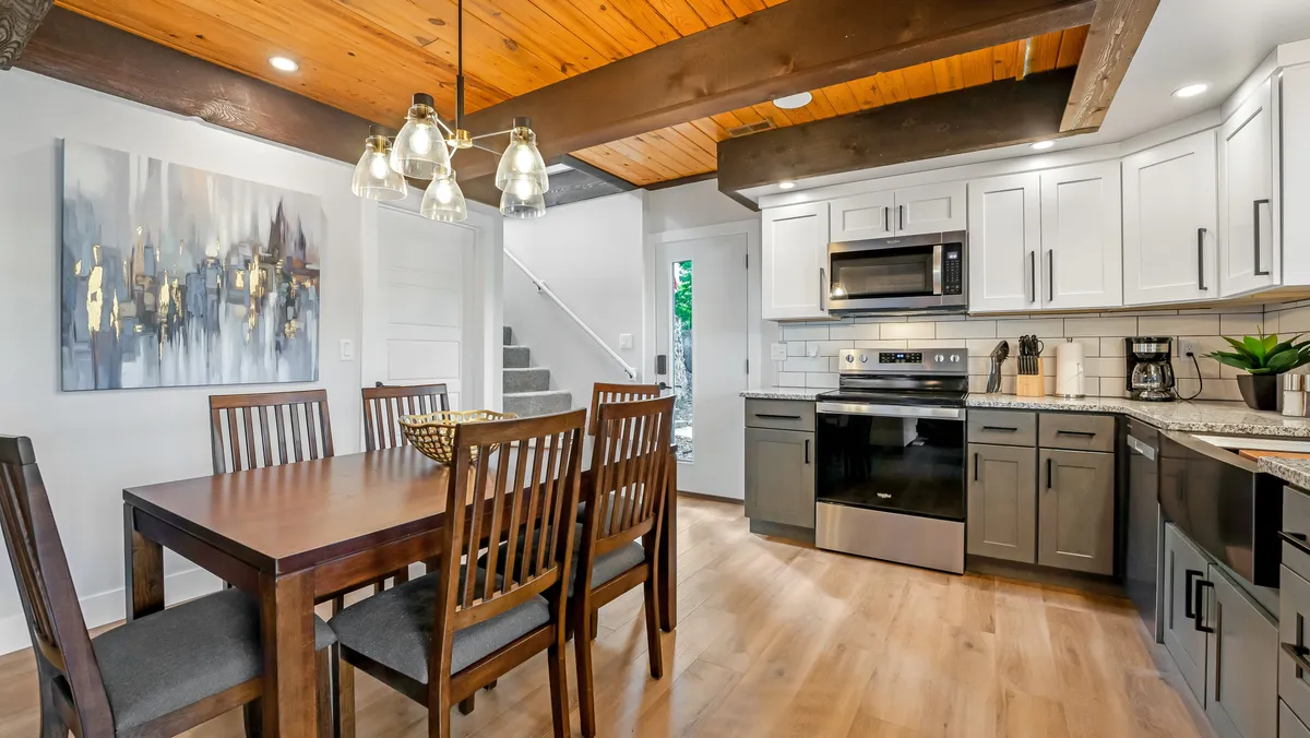 The image features a spacious dining table adjacent to kitchen appliances, with artwork on the wall and a visible staircase. The setting feels warm and welcoming, ideal for shared meals, celebrations, and social gatherings.