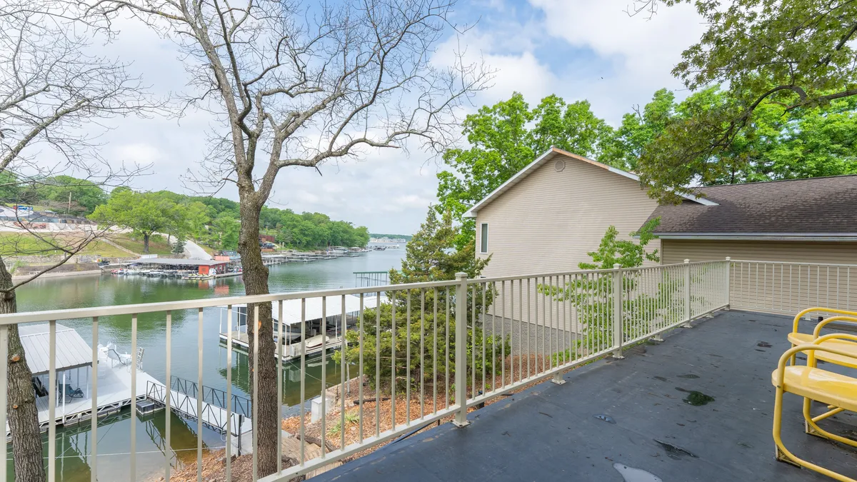 This image presents a sweeping lake view from a vantage point near a dock, featuring tranquil water, the dock's structure, adjacent greenery, and a balcony railing in the foreground. The mood is peaceful, inviting viewers to imagine themselves relaxing or preparing for a day of lake activities right at the water's edge.