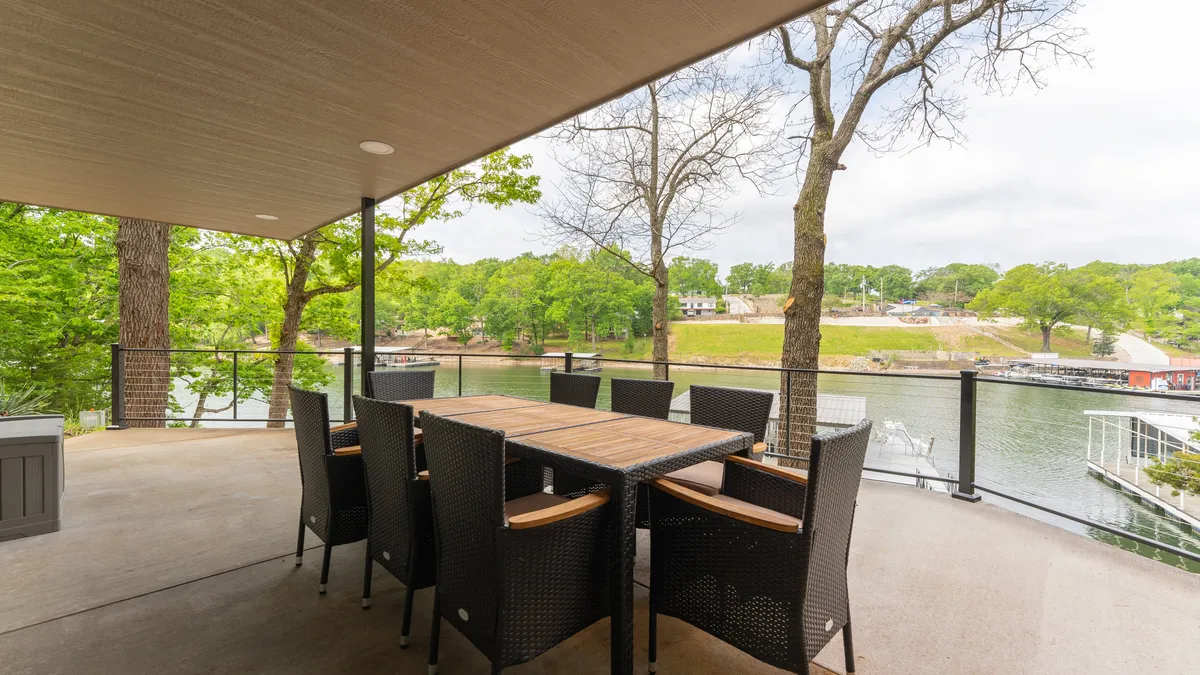 This image features an outdoor dining set on an expansive deck overlooking the lake, surrounded by lush greenery. The setup is perfect for enjoying meals outdoors, relaxing with friends, or taking in serene water views. The spacious deck area provides a seamless blend of sun and shade, inviting guests to lounge, dine, or socialize just steps from the water.
